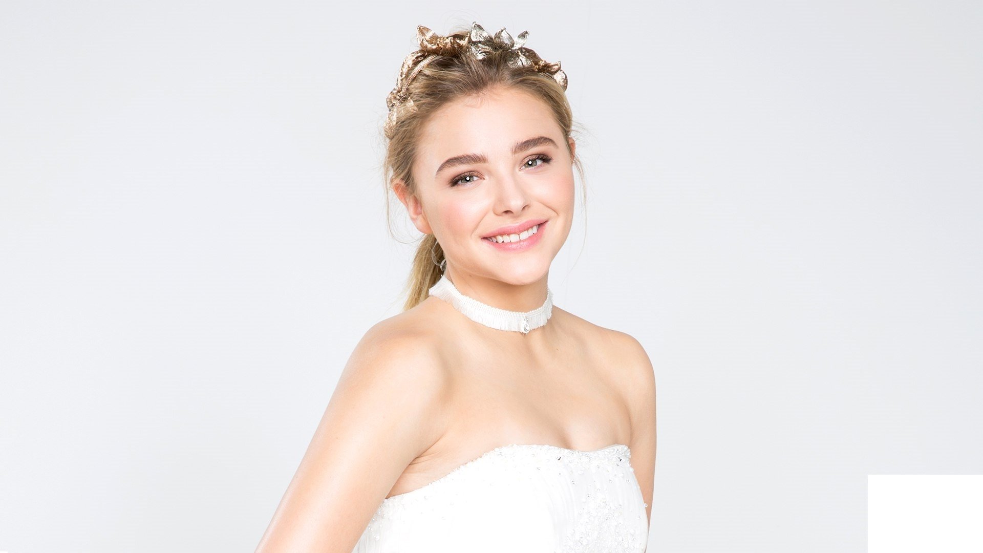 A smiling Chloë Grace Moretz, a blonde American actress, poses gracefully in a light outfit against a clean white background, creating an inviting HD desktop wallpaper.