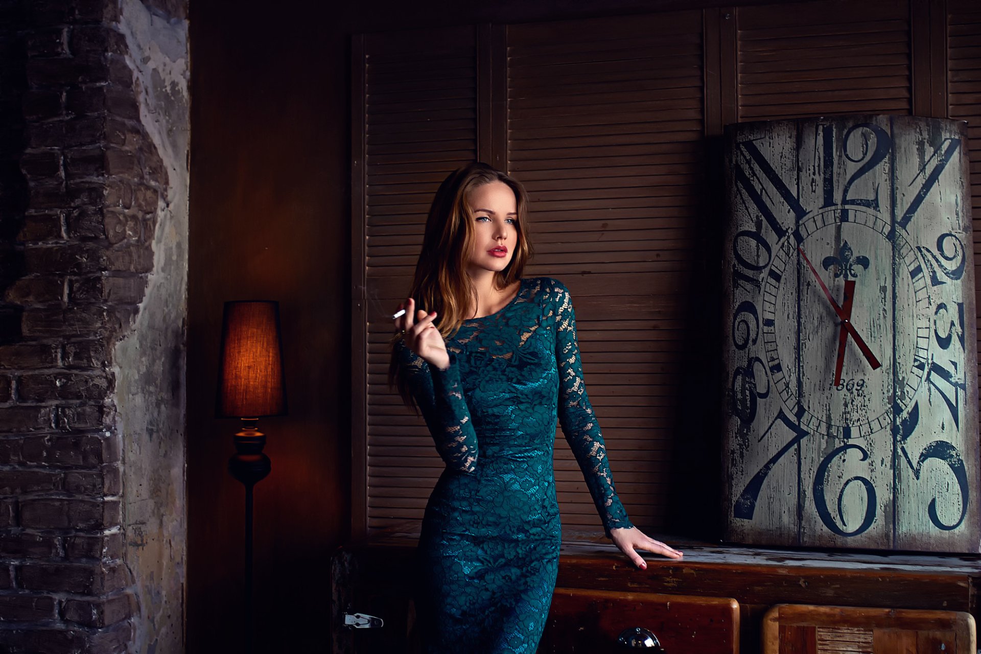 Brunette woman with blue eyes and red lipstick in a teal dress, smoking while standing in front of a large vintage clock. Background features dim lighting and a rustic atmosphere. HD desktop wallpaper.