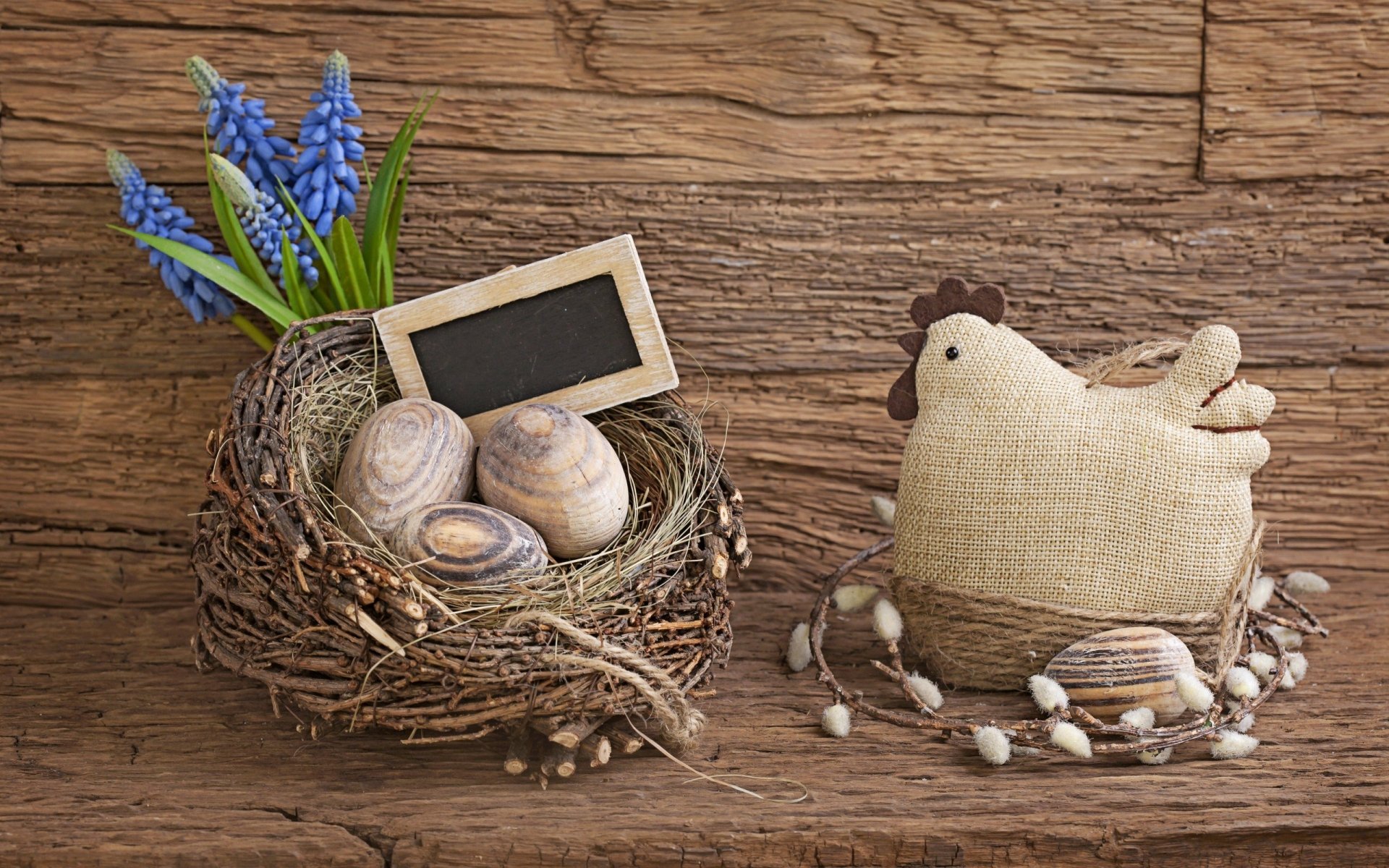 A charming HD desktop wallpaper of an Easter nest with eggs, a small chalkboard, blue flowers, and a fabric chicken on a rustic wooden background.