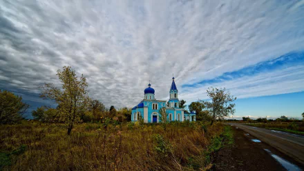 HD desktop wallpaper of a blue chapel with religious architecture set against a dramatic cloudy sky, surrounded by autumnal grass and a nearby road.