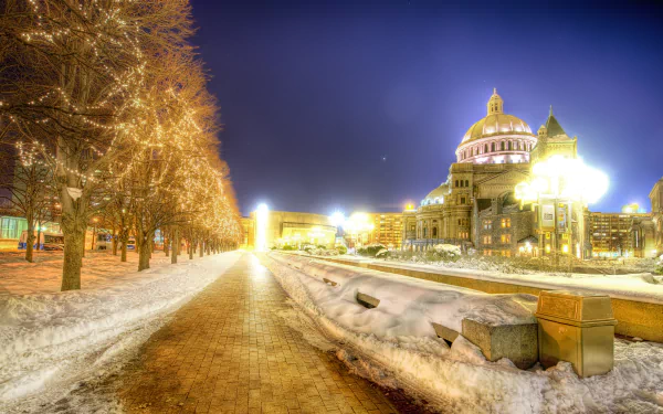 A winter night scene in Boston featuring a snowy walkway lined with Christmas lights, leading to a beautifully lit building under a dark blue sky.