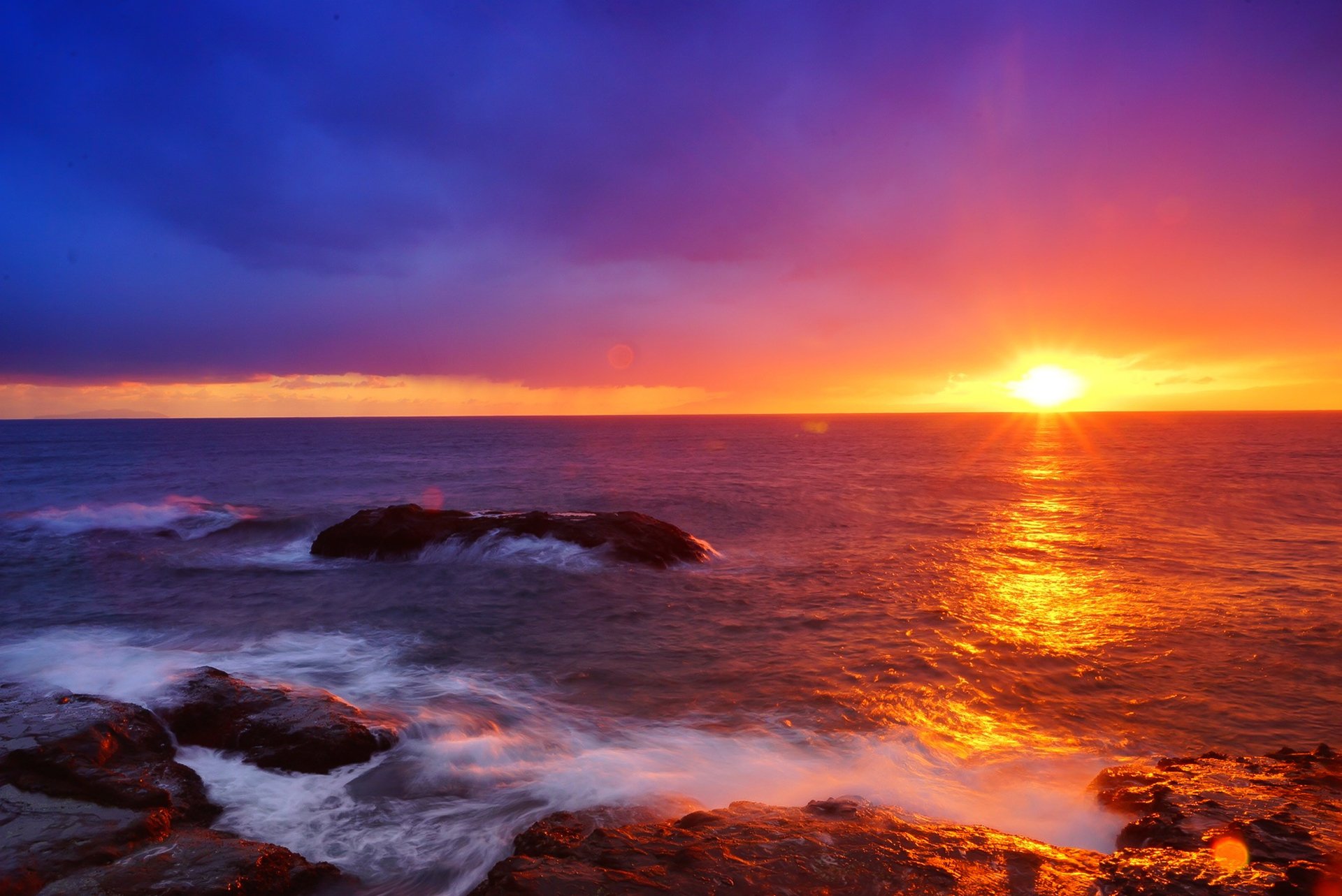 HD PC desktop wallpaper: sunrise on the ocean horizon, golden sun reflecting on waves and rocky shore beneath a vivid sunset-colored sky — serene nature scene.