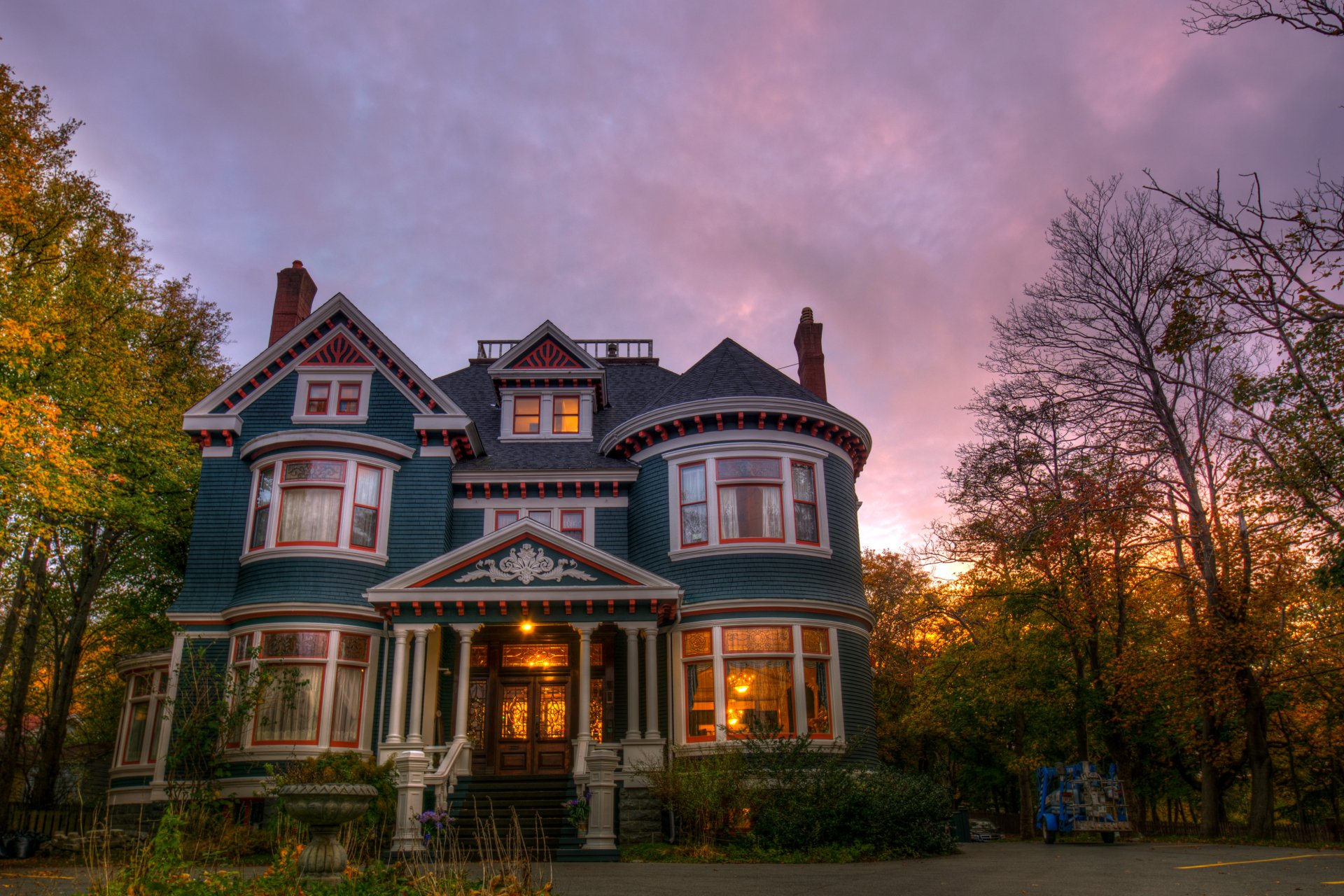 2K Quad HD PC desktop wallpaper and background: lighted, man-made Victorian house at dusk — ornate blue home with warm interior lights, trees and a pink sky.