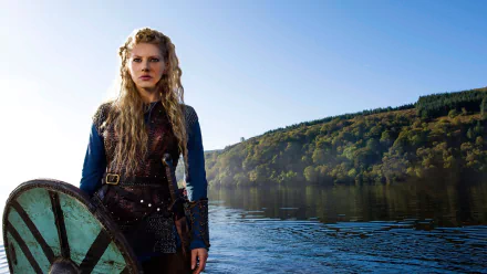 Lagertha (Vikings, 2013) in armor with shield standing at a calm lakeside under a clear blue sky — HD PC desktop wallpaper background.
