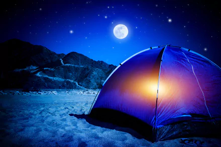 A vibrant tent glows under a starry sky, with a bright moon illuminating the night. Sand surrounds the campsite, framed by distant mountains, creating a serene camping atmosphere.