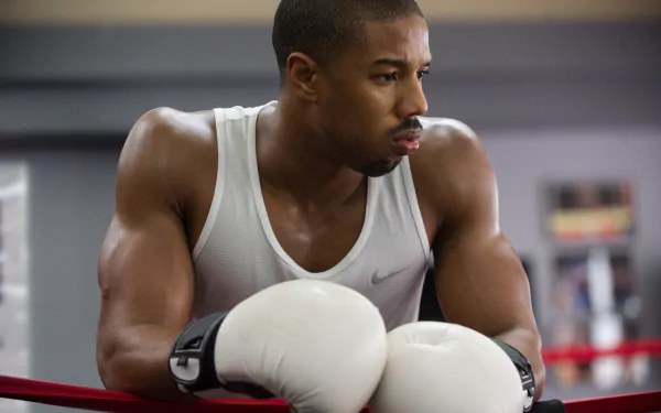 4K movie desktop wallpaper: muscular boxer in a white tank top and gloves leaning on ring ropes, focused and pensive between rounds.