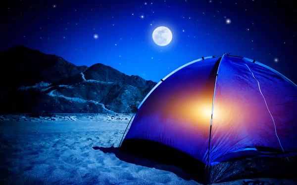 A vibrant tent glows under a starry sky, with a bright moon illuminating the night. Sand surrounds the campsite, framed by distant mountains, creating a serene camping atmosphere.