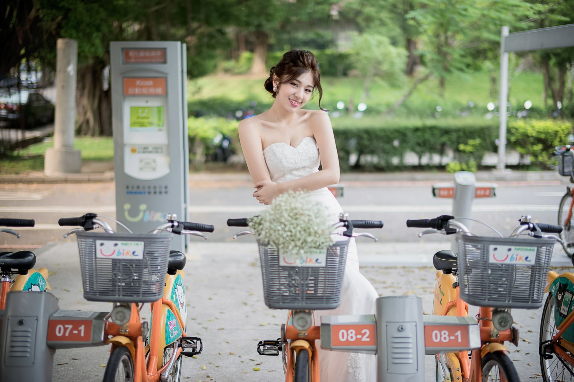 A smiling brunette bride in a wedding dress stands among a row of bicycles in a scenic outdoor setting, with greenery in the background.