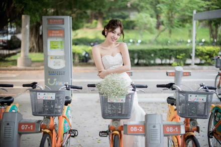 A smiling brunette bride in a wedding dress stands among a row of bicycles in a scenic outdoor setting, with greenery in the background.