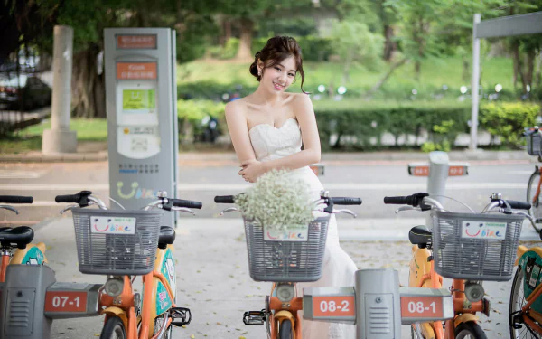 A smiling brunette bride in a wedding dress stands among a row of bicycles in a scenic outdoor setting, with greenery in the background.