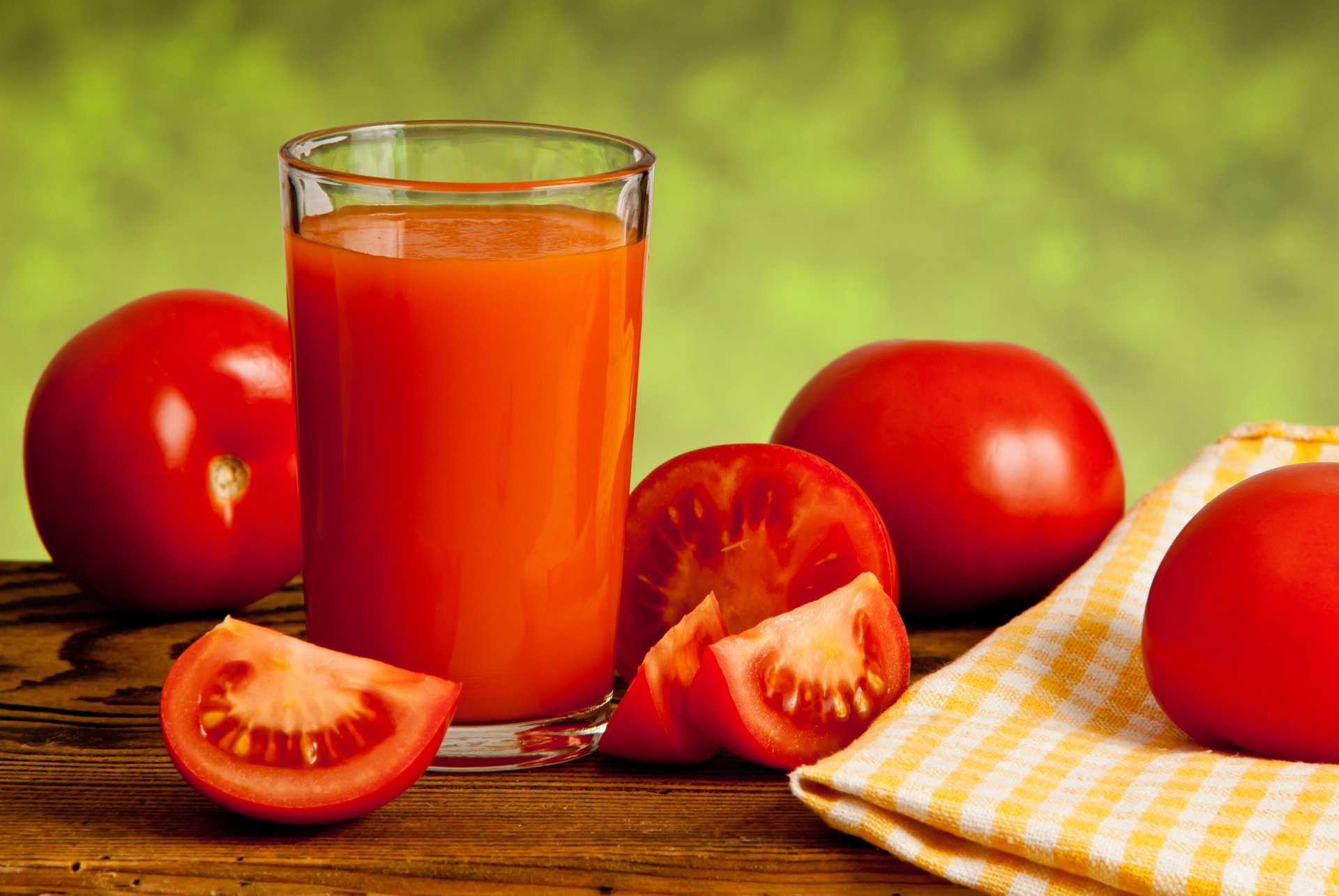 A glass of vibrant red tomato juice surrounded by fresh whole and sliced tomatoes on a wooden surface with a yellow checkered cloth, captured in 8K Ultra HD.