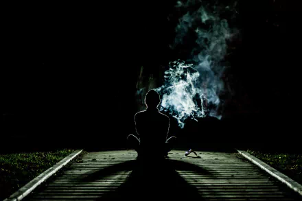 A serene silhouette of a person meditating alone on a wooden path, surrounded by smoke against a black night, creating a spiritual and tranquil atmosphere for an HD desktop wallpaper.