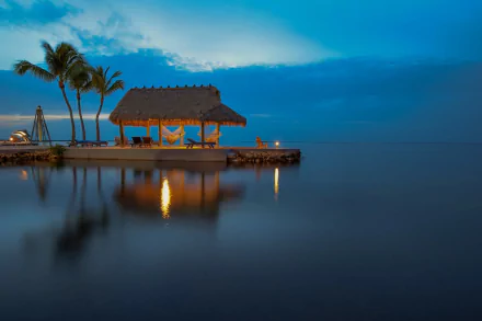 A tranquil tropical scene at dusk, featuring a thatched bungalow beside the calm ocean, with palm trees, a hammock, and gentle reflections, creating a serene holiday atmosphere.