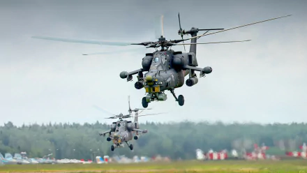 HD desktop wallpaper featuring two Mil Mi-28 military helicopters flying low over a runway with a cloudy sky and forest in the background.