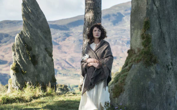 Claire Fraser, portrayed by Caitriona Balfe in the TV show Outlander, stands gracefully amidst ancient stones, with a scenic landscape in the background, showcasing a moment of connection to her surroundings.