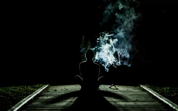 A serene silhouette of a person meditating alone on a wooden path, surrounded by smoke against a black night, creating a spiritual and tranquil atmosphere for an HD desktop wallpaper.