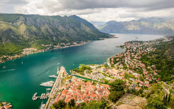 Aerial view of Kotor, Montenegro, showcasing coastal houses, boats docked by the marina, and the mountainous cityscape along the bay.