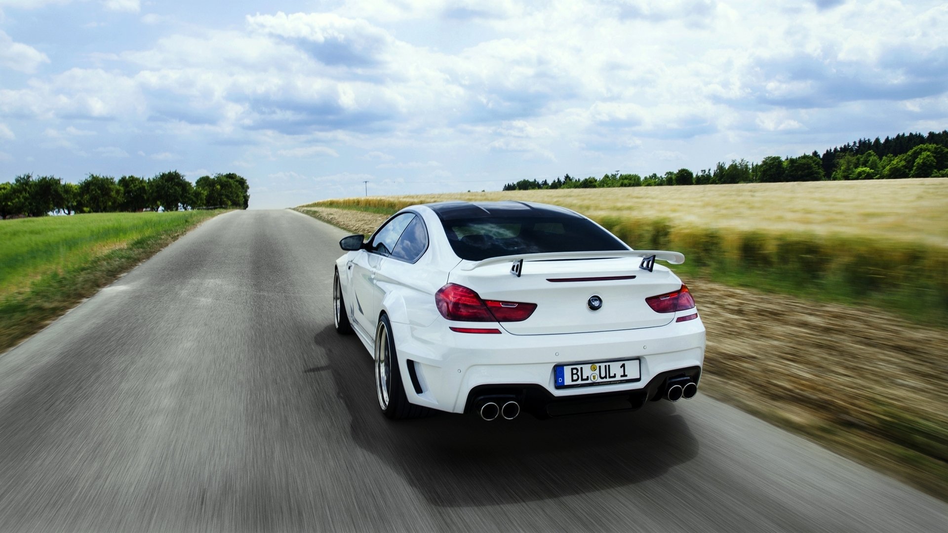 White BMW M6 coupe seen from the rear three-quarter angle driving on a country road, presented as a 2K Quad HD PC desktop wallpaper/background.
