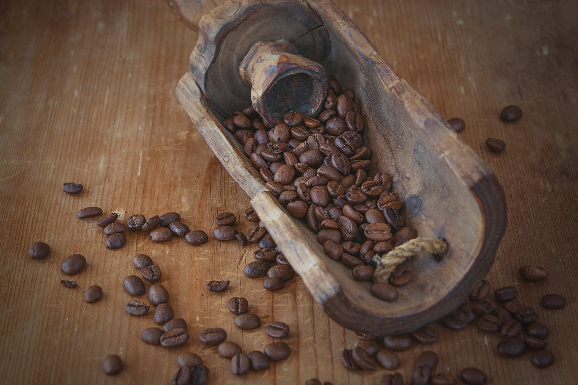 A rustic wooden scoop filled with brown coffee beans rests on a wooden surface, capturing the essence of coffee in this HD desktop wallpaper.