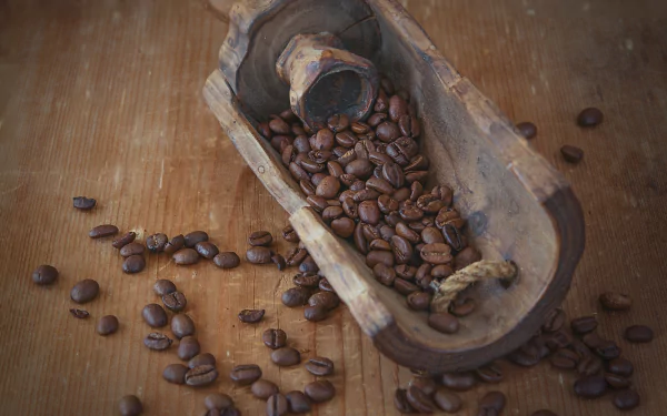 A rustic wooden scoop filled with brown coffee beans rests on a wooden surface, capturing the essence of coffee in this HD desktop wallpaper.