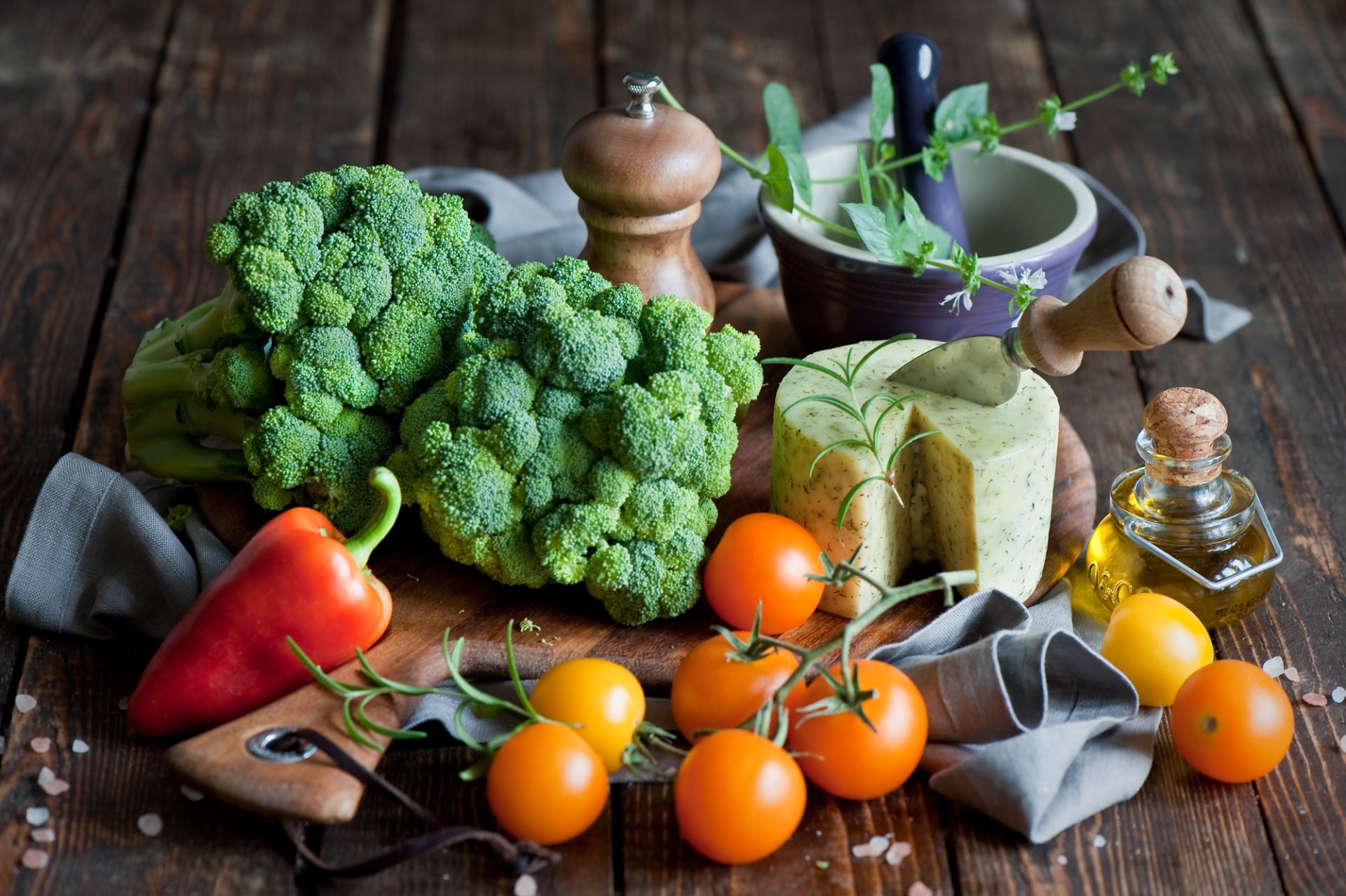 A still life HD desktop wallpaper featuring fresh broccoli, cherry tomatoes, red pepper, cheese, olive oil, and herbs on a rustic wooden surface.