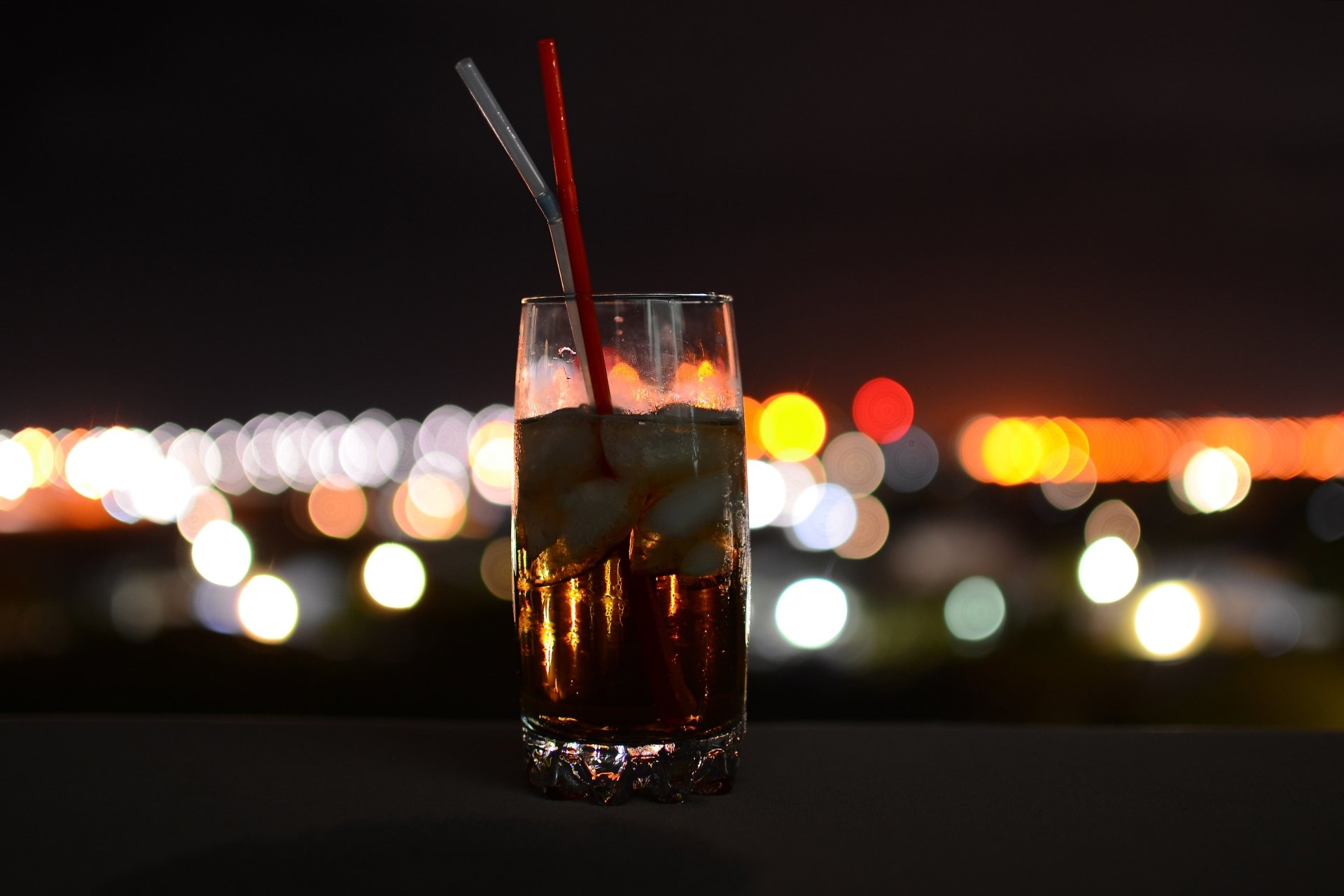 4K Ultra HD PC desktop background of a glass of Coca‑Cola with ice and two straws, a food/drink shot framed against colorful bokeh city lights.