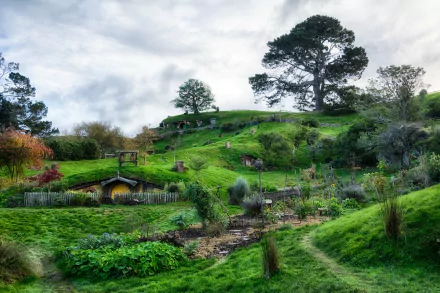 HD desktop wallpaper of the man-made Hobbiton set featuring lush green hills, cozy hobbit holes, and vibrant gardens under a cloudy sky.