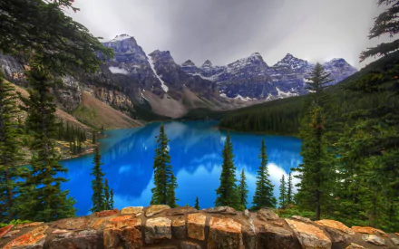 HD desktop wallpaper of Moraine Lake in Canada showcasing vibrant blue water, surrounded by evergreen trees and towering mountain peaks under a cloudy sky.