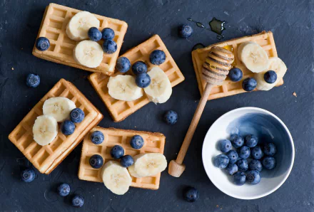HD desktop wallpaper featuring a still life breakfast spread with waffles topped with banana slices and blueberries, accompanied by a honey dipper and a bowl of fresh blueberries.
