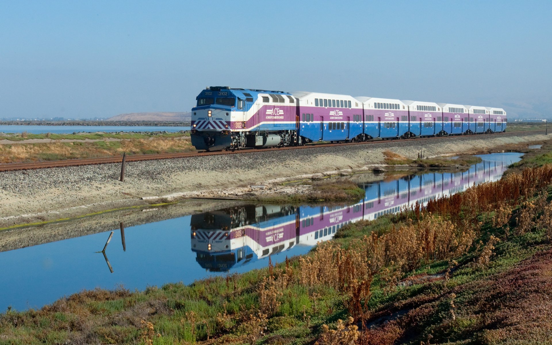 HD desktop wallpaper showing a passenger train traveling alongside a calm waterway, with the train and landscape reflected in the water.