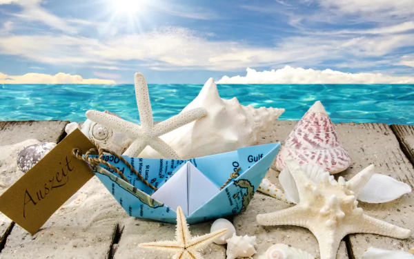Artistic 4K Ultra HD desktop wallpaper featuring a paper boat made from a map, surrounded by various seashells and starfish on a sandy beach with a summer ocean backdrop.
