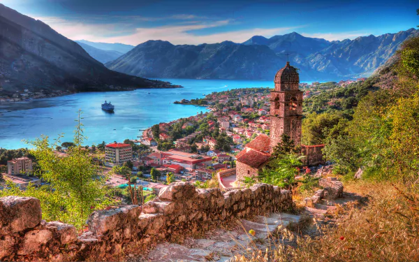 2K Quad HD PC desktop wallpaper: panoramic Kotor, Montenegro—mountain-ringed bay with a ship, terracotta-roofed town, man-made stone church tower and houses framed by rocky foreground.