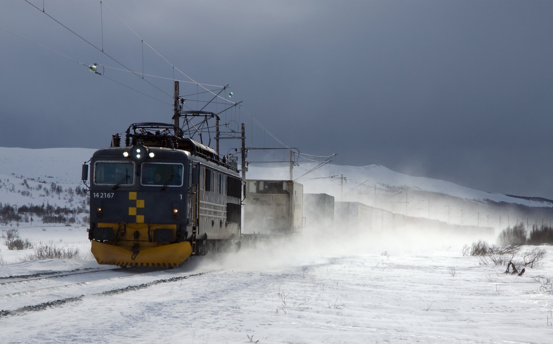 A powerful train travels through a snowy landscape, creating a cloud of snow as it moves along the tracks under a moody sky. A striking HD desktop wallpaper for enthusiasts of vehicles and trains.