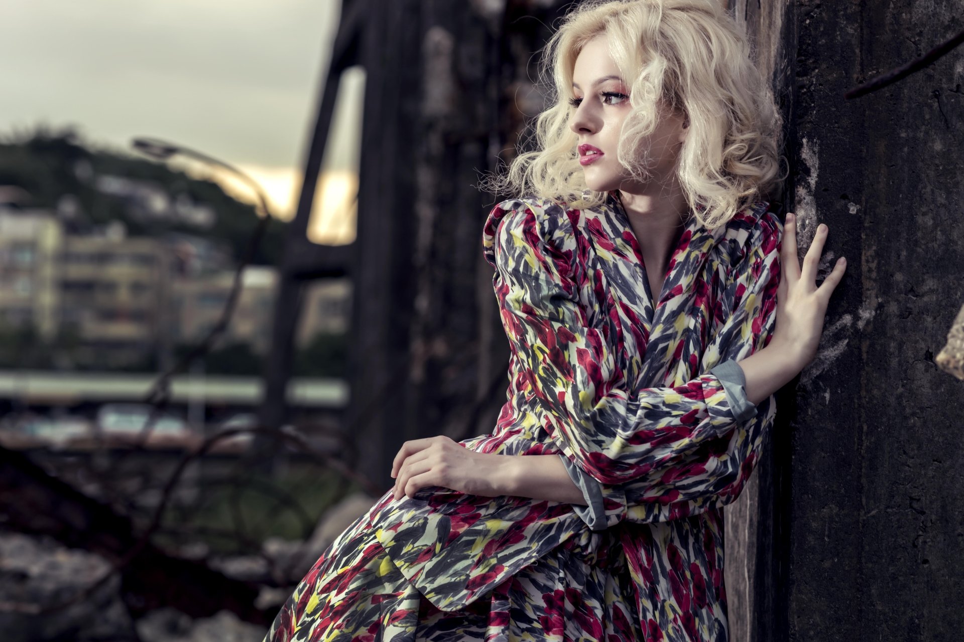 Blonde female model in patterned dress leans against a wall with blurred urban background — 8K Ultra HD PC desktop wallpaper/background