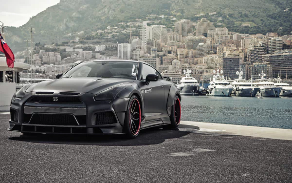 A sleek silver Nissan GT-R parked along a scenic waterfront, showcasing its dynamic design against a backdrop of luxurious yachts and a mountainous cityscape.