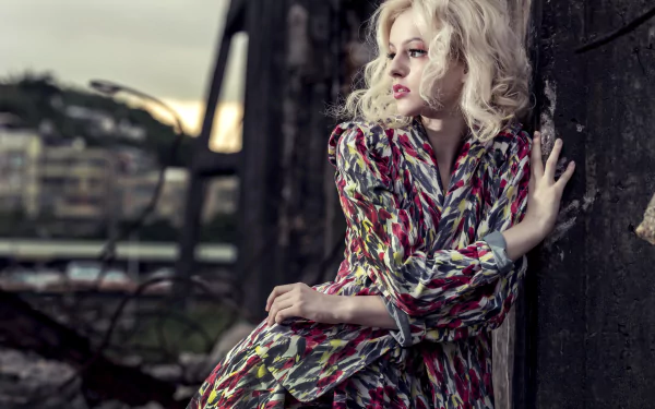 Blonde female model in patterned dress leans against a wall with blurred urban background — 8K Ultra HD PC desktop wallpaper/background