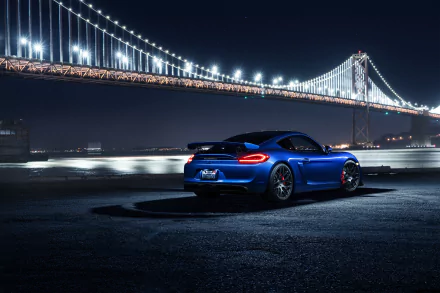 A blue Porsche 718 Cayman car parked near a waterfront at night, with a brightly lit bridge in the background, featured as an HD PC desktop wallpaper.