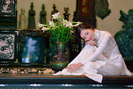 A Vietnamese woman wearing a traditional white Ao Dai sits gracefully near a vase of lilies in an ornate setting, captured in 4K Ultra HD.