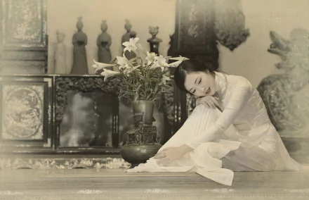 A serene Vietnamese woman in traditional Ao Dai sits gracefully beside a vase of lilies, captured in a soft sepia tone, evoking elegance and cultural beauty.