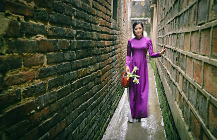 Vietnamese woman model in a vibrant purple Áo Dài holding a lily, walking through a narrow alley with brick walls in this 4K Ultra HD desktop wallpaper.