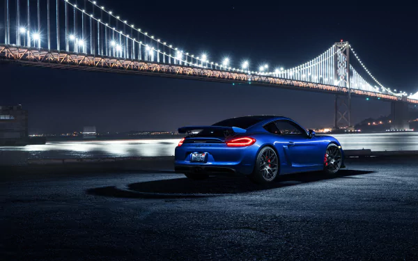 A blue Porsche 718 Cayman car parked near a waterfront at night, with a brightly lit bridge in the background, featured as an HD PC desktop wallpaper.
