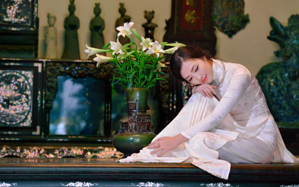 A Vietnamese woman wearing a traditional white Ao Dai sits gracefully near a vase of lilies in an ornate setting, captured in 4K Ultra HD.