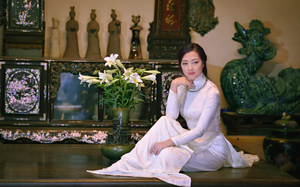 A Vietnamese woman wearing a traditional Ao Dai poses elegantly beside a vase of lilies, set against a backdrop of ornate decor, showcasing cultural beauty in this 4K Ultra HD wallpaper.