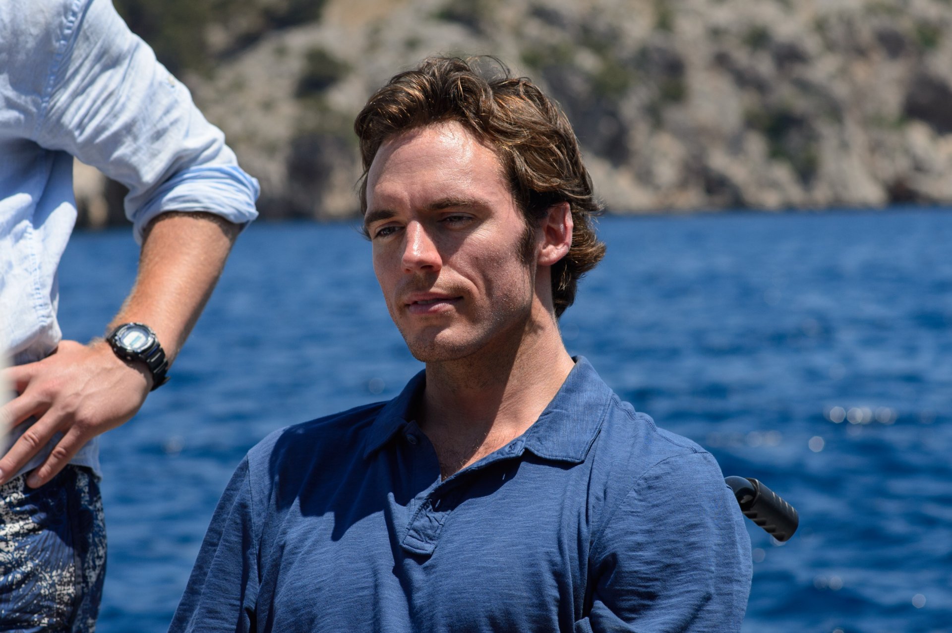 HD desktop wallpaper featuring Sam Claflin in a scene from the movie Me Before You, set against a backdrop of blue water and rocky cliffs.