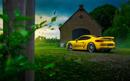 A vibrant yellow Porsche Cayman GT4 parked on a gravel path near a rustic stone building, set against a lush green landscape under a moody sky.
