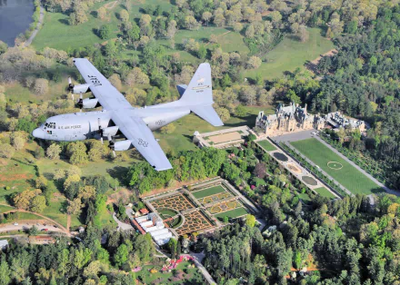 A Lockheed C-130 Hercules military transport aircraft flies over lush greenery and formal gardens, captured in high-definition as a desktop wallpaper.