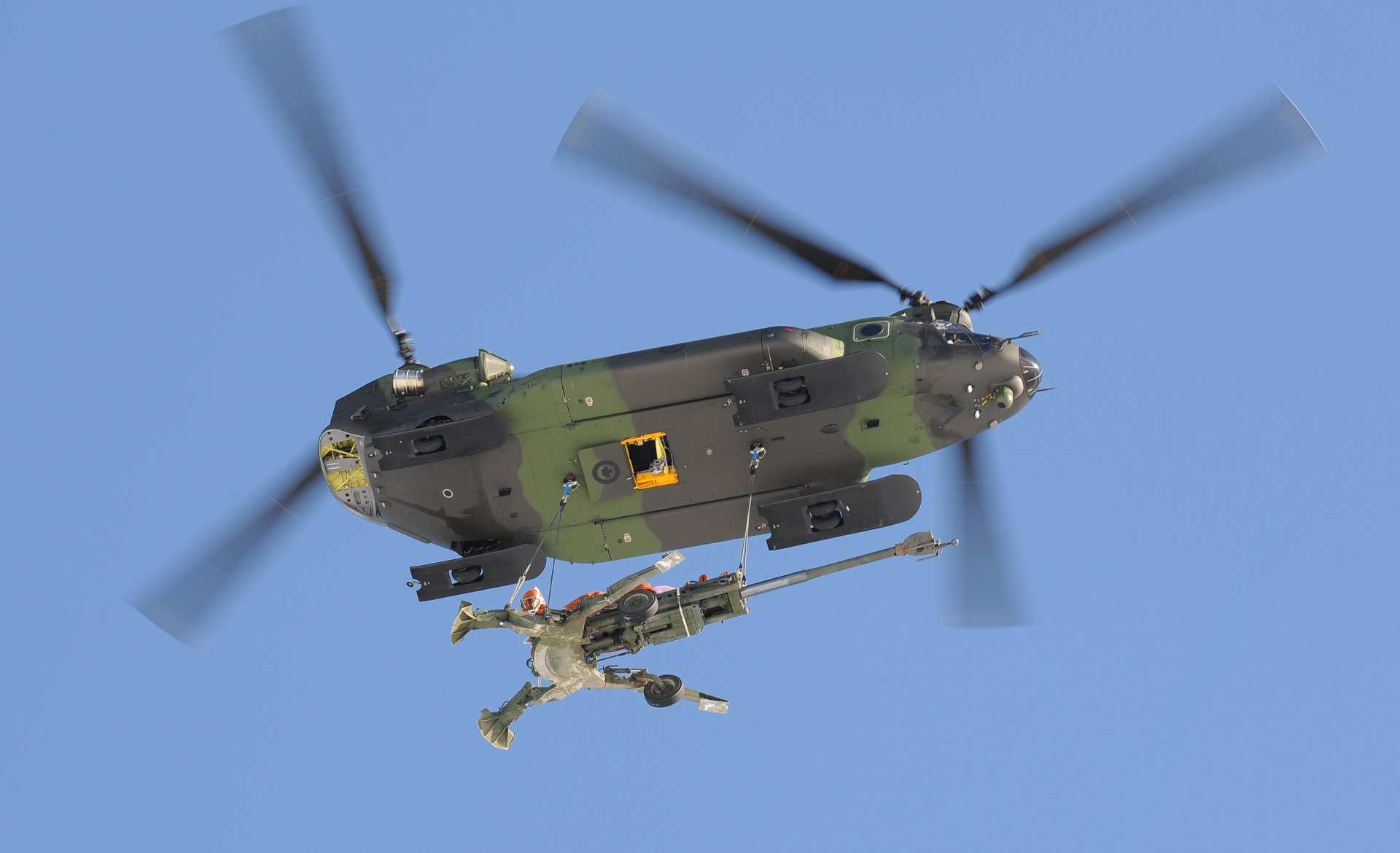 Boeing CH-47 Chinook helicopter flying against a clear blue sky, transporting a military Howitzer M777 artillery piece.