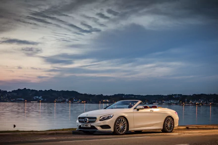 A sleek white Mercedes-Benz S-Class convertible parked by the waterfront, showcasing its elegance against a vibrant sunset sky. Perfect 4K Ultra HD desktop wallpaper.