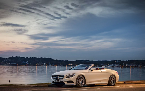 A sleek white Mercedes-Benz S-Class convertible parked by the waterfront, showcasing its elegance against a vibrant sunset sky. Perfect 4K Ultra HD desktop wallpaper.