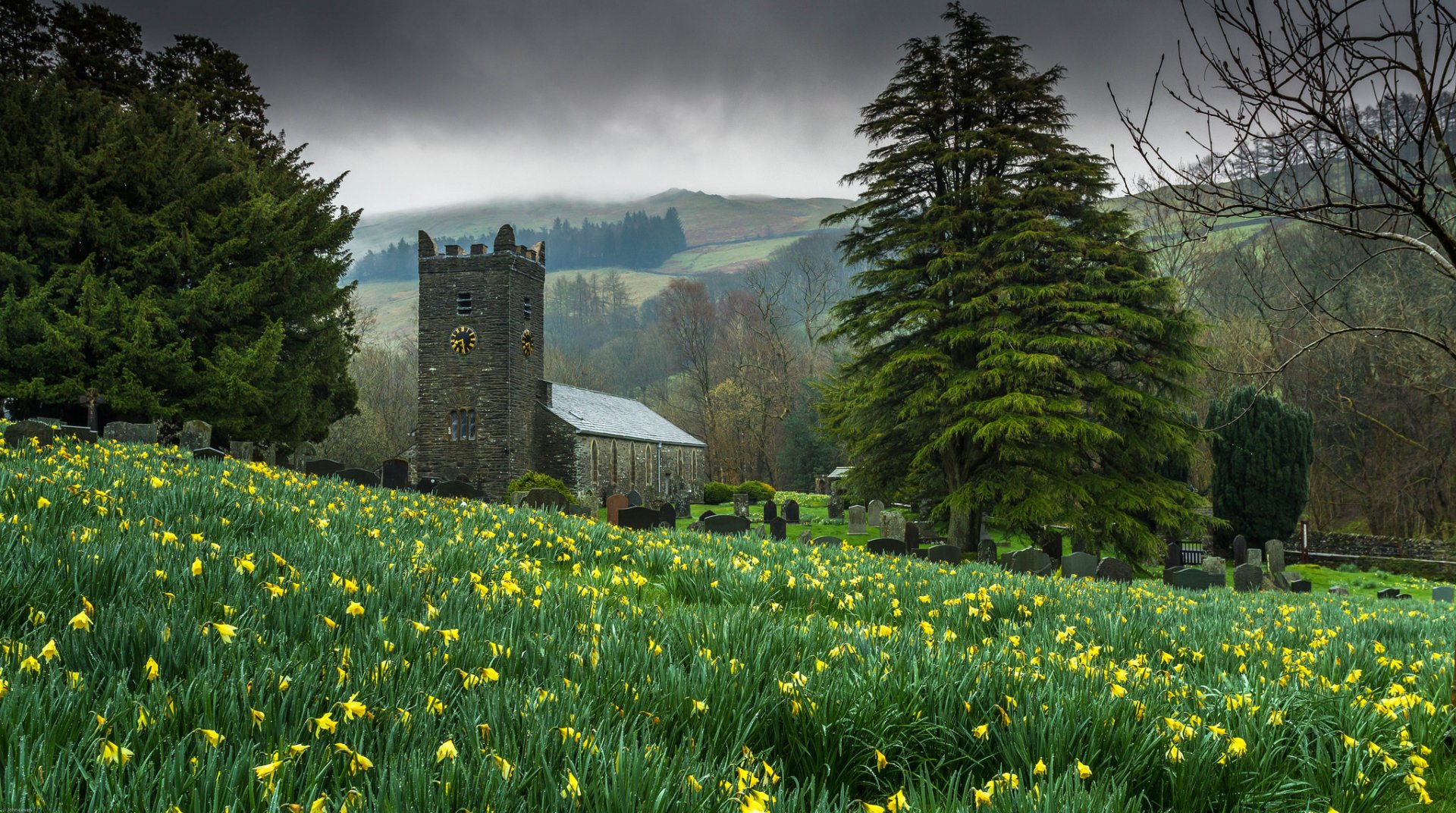 HD desktop wallpaper featuring a serene country graveyard with blooming flowers, tall trees, and a historic church under a cloudy sky.
